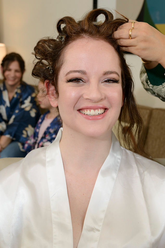 A radiant bride smiling joyfully while getting her hair styled on her wedding day. She is wearing a silky white robe, with curls set in clips and makeup complete. Bridesmaids in floral robes can be seen in the background, sharing the joyful moment.