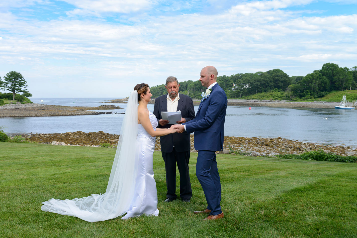 A serene wedding ceremony by the ocean, with the radiant bride outdoors in a stunning white gown with elegant draping and groom in a blue suit holding hands and exchanging vows under the guidance of the officiant, framed by lush greenery and a tranquil shoreline in the background.