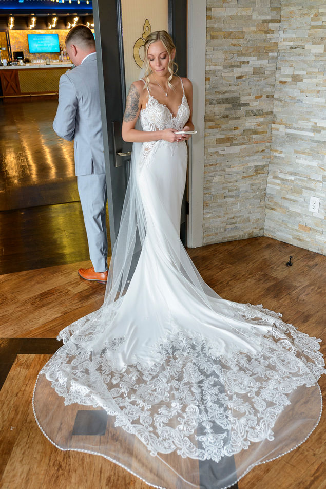 Bride in a stunning white wedding gown with a long, intricate lace train, standing by a doorway and holding vows while looking down. A groom in a gray suit stands behind the door, facing away, creating an emotional pre-ceremony moment.
