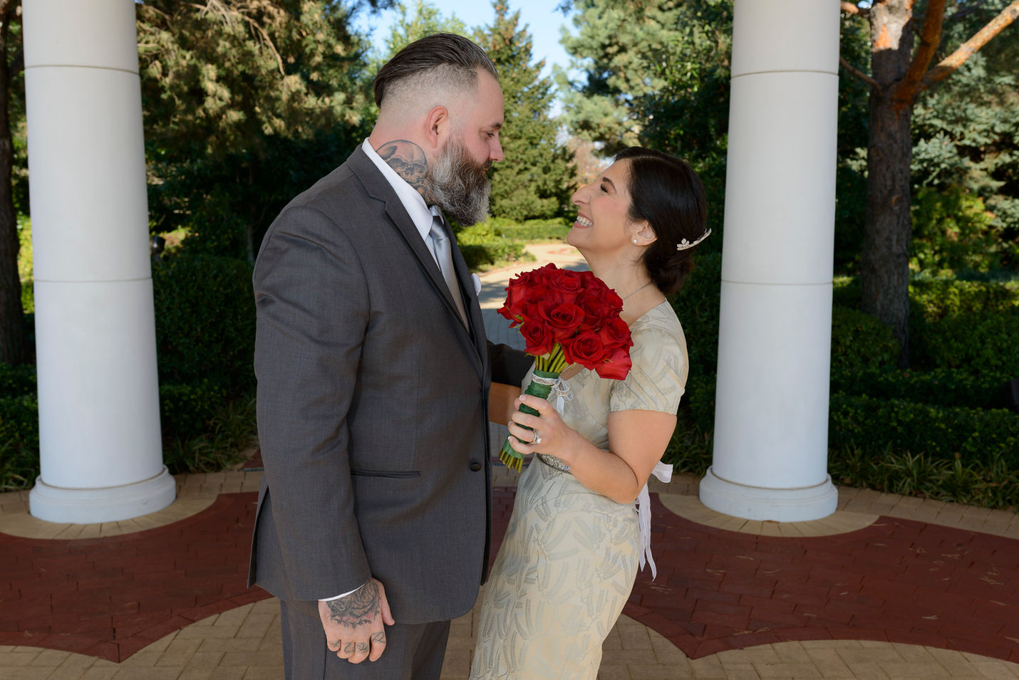 A joyful bride and groom share a loving moment after "first look" on their wedding day. The bride, dressed in an elegant champagne-colored gown and holding a vibrant bouquet of red roses, smiles warmly at the groom. The groom, wearing a sharp gray suit with a tattoo visible on his neck and hand, gazes back at her with affection. They stand under a columned structure, surrounded by lush greenery and natural light.