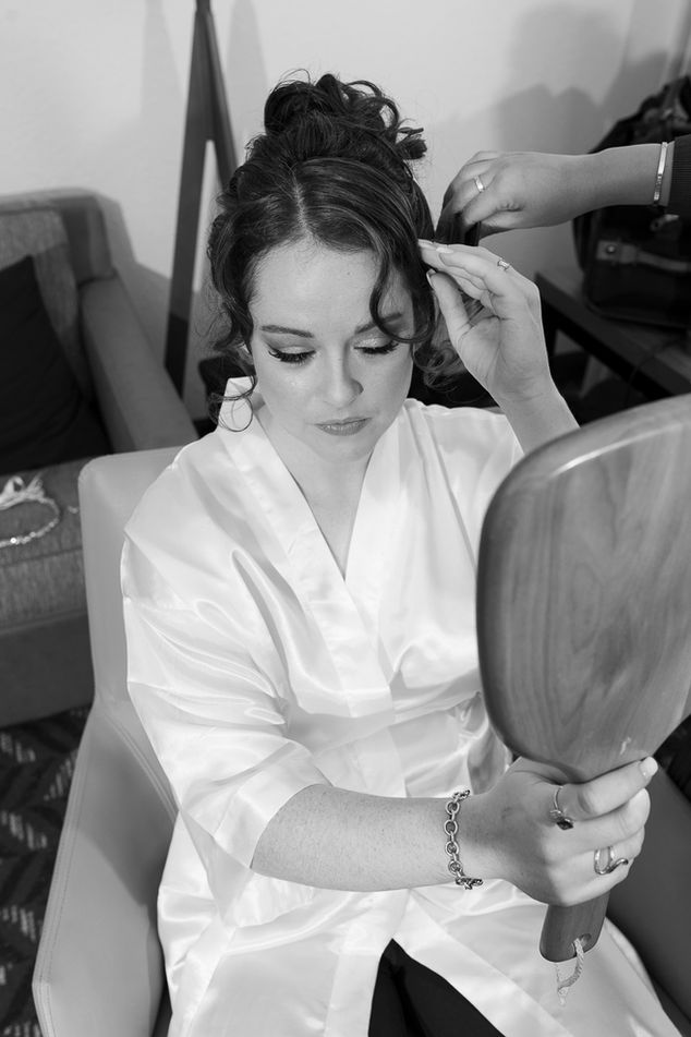 Black and white photograph of a bride sitting in a chair, wearing a satin robe as she adjusts her hair. She holds a wooden hand mirror in one hand while a stylist works on her updo in the background. The bride's face shows concentration and grace.