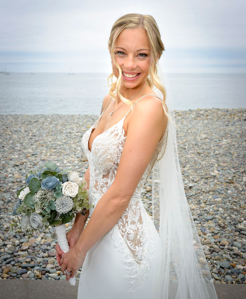 A radiant bride stands on a pebble beach, holding a bouquet of blue and white flowers. She smiles brightly, dressed in an elegant lace wedding gown with delicate straps and a long flowing veil. The calm ocean provides a serene backdrop, enhancing the beaming
expression on her face.
