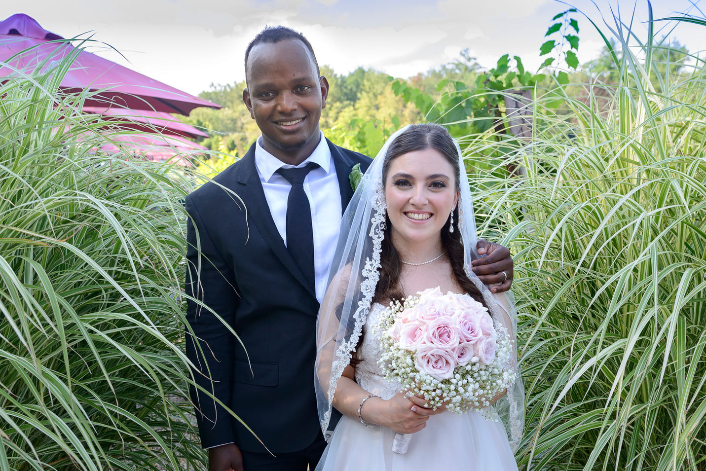 A just-married couple poses for a portrait outdoors, radiating happiness. The groom wears a black suit with a white shirt and tie, while the bride shines in a white lace gown with a veil, holding a bouquet of pink roses and baby's breath. They stand close together, framed by vibrant greenery and soft natural light