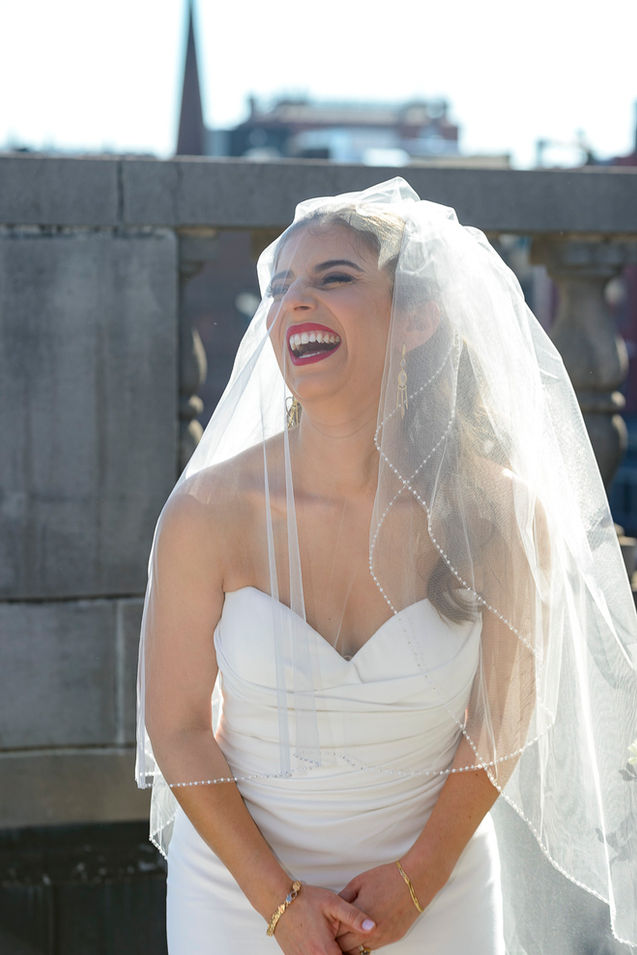 The bride stands outdoors at the rooftop ceremony, laughing joyfully under a delicate, pearl-lined veil. She wears a strapless white gown and gold jewelry, with sunlight illuminating her face and veil. A blurred cityscape provides a scenic backdrop.