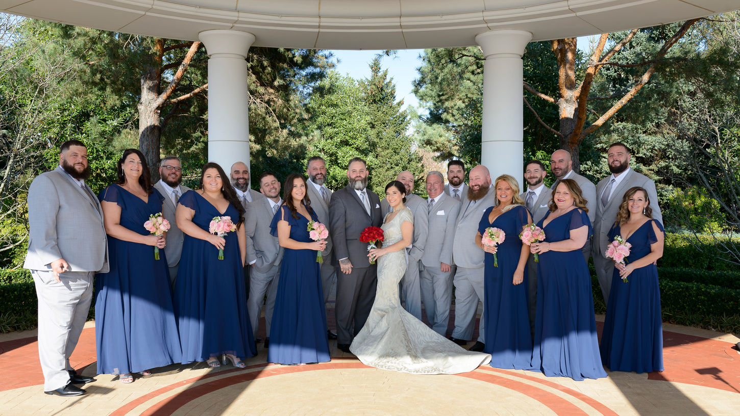 A wedding group photograph taken under a large gazebo. The bride and groom stand in the center, with the bride wearing an elegant ivory gown with a long train and holding a bouquet of red roses. The groom is dressed in a dark gray suit with a light gray tie. Surrounding them are the wedding party: bridesmaids in deep blue dresses holding pastel pink bouquets and groomsmen in light gray suits. The backdrop features lush greenery and tall trees, creating a serene outdoor setting for the celebration.