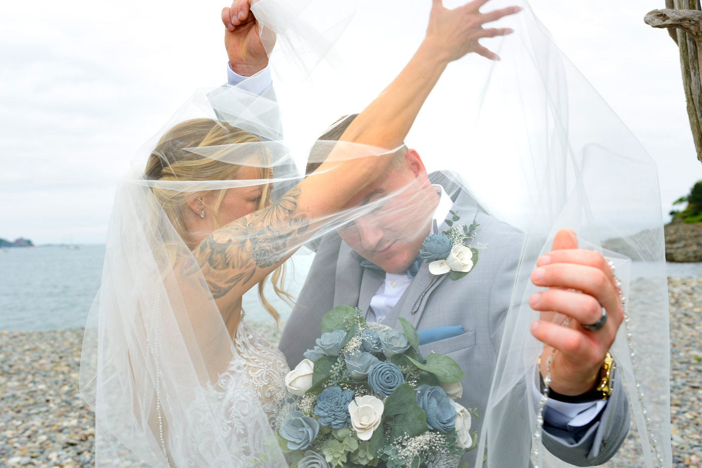 Bride and groom playfully adjust the bride's veil together at the beach, creating a dynamic and intimate moment. The groom, dressed in a light gray suit with a blue tie and floral boutonniere, slightly bends forward under the veil, while the bride, showcasing a detailed lace gown and a tattooed arm, laughs as she helps to lift the veil. The backdrop features a pebble beach and a distant ocean horizon.