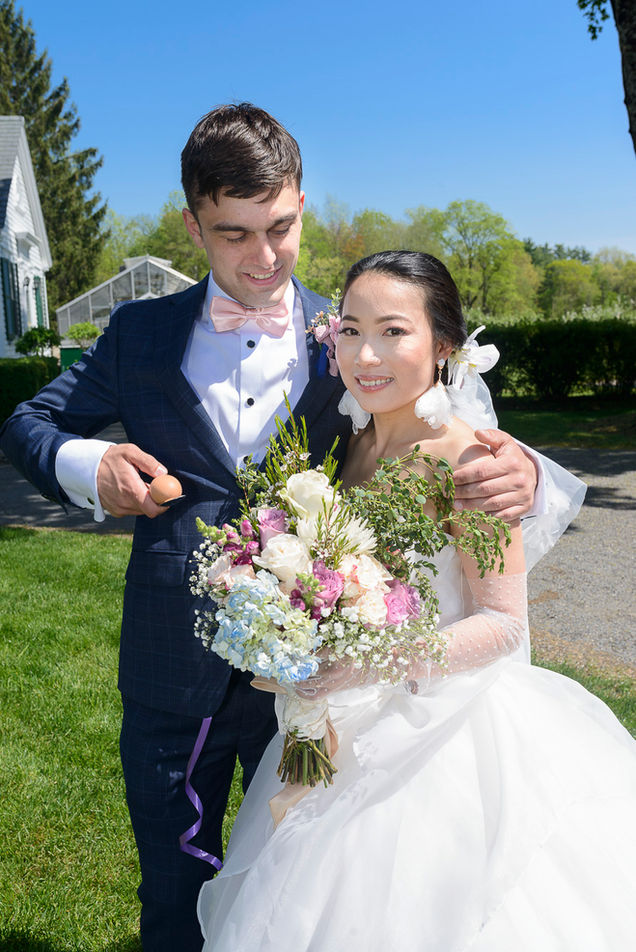 Newlywed couple smiling joyfully in an outdoor garden setting under a bright blue sky. The groom, dressed in a navy suit with a pink bow tie, playfully holds an egg in his hand. The bride, wearing a white wedding gown with polka-dot sleeves, holds a vibrant bouquet of mixed flowers, including roses, hydrangeas, and baby's breath, accented with greenery.