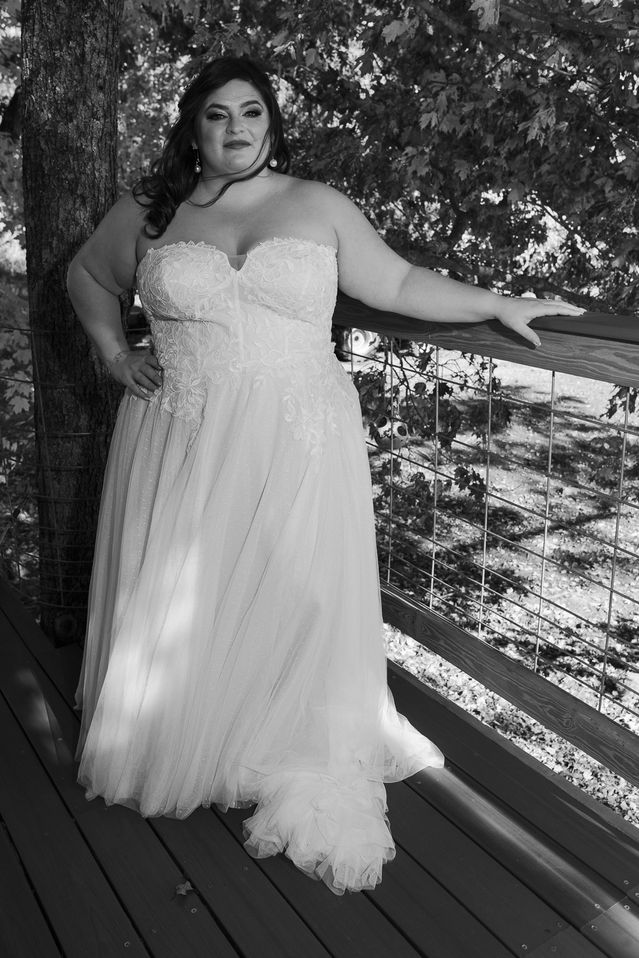 A bride poses confidently on a wooden deck in a strapless lace wedding gown, her hand resting on the railing with autumn leaves providing a picturesque backdrop in this black-and-white photograph.