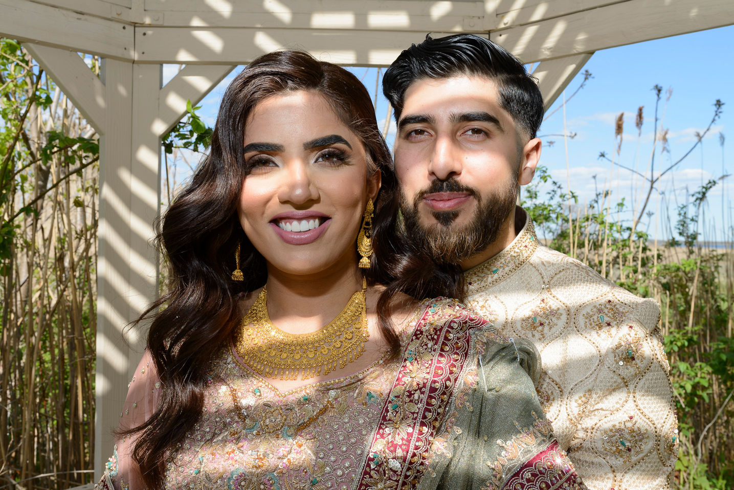 A closeup portrait of a couple during a Pakistani wedding celebration. The bride is adorned in an intricately embroidered dress featuring gold and maroon accents, complemented by gold jewelry and cascading curls. The groom stands beside her in a cream and gold sherwani, with an affectionate expression. The couple is framed by natural light and shadows cast by a wooden pergola, with reeds and a bright blue sky visible in the background.