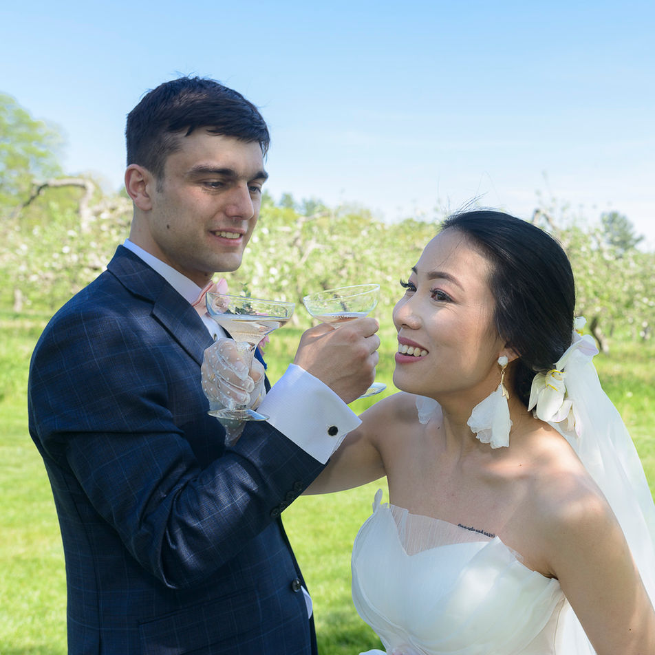 Bride and groom sharing a toast with champagne in an outdoor apple blossom orchard setting. The groom, wearing a navy suit and pink bow tie, gazes at the bride, who smiles brightly while holding her glass. The bride is dressed in a strapless white gown with floral earrings, her veil adorned with orchids, creating a romantic and celebratory moment against the lush greenery and blue sky.