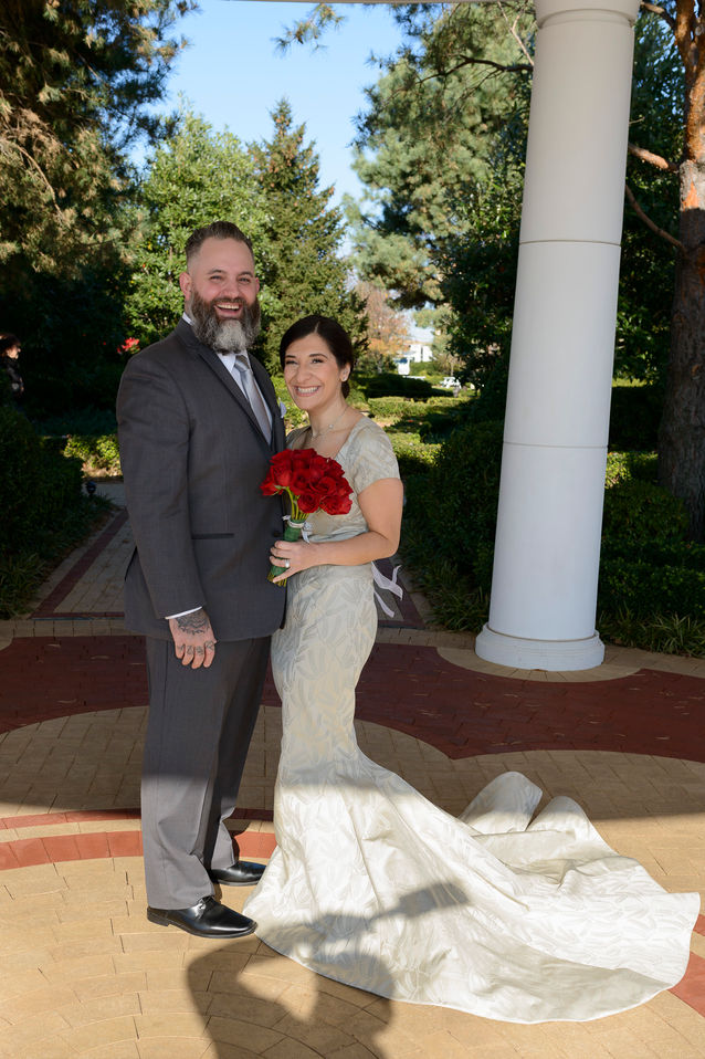 A beautiful wedding photograph featuring a smiling bride and groom standing together outdoors. The bride wears an elegant ivory gown with a long train, holding a bouquet of vibrant red roses, while the groom is dressed in a dark gray suit with a light gray tie. They are standing under a white columned structure, surrounded by lush greenery and trees on a sunny day. The scene exudes happiness and love.