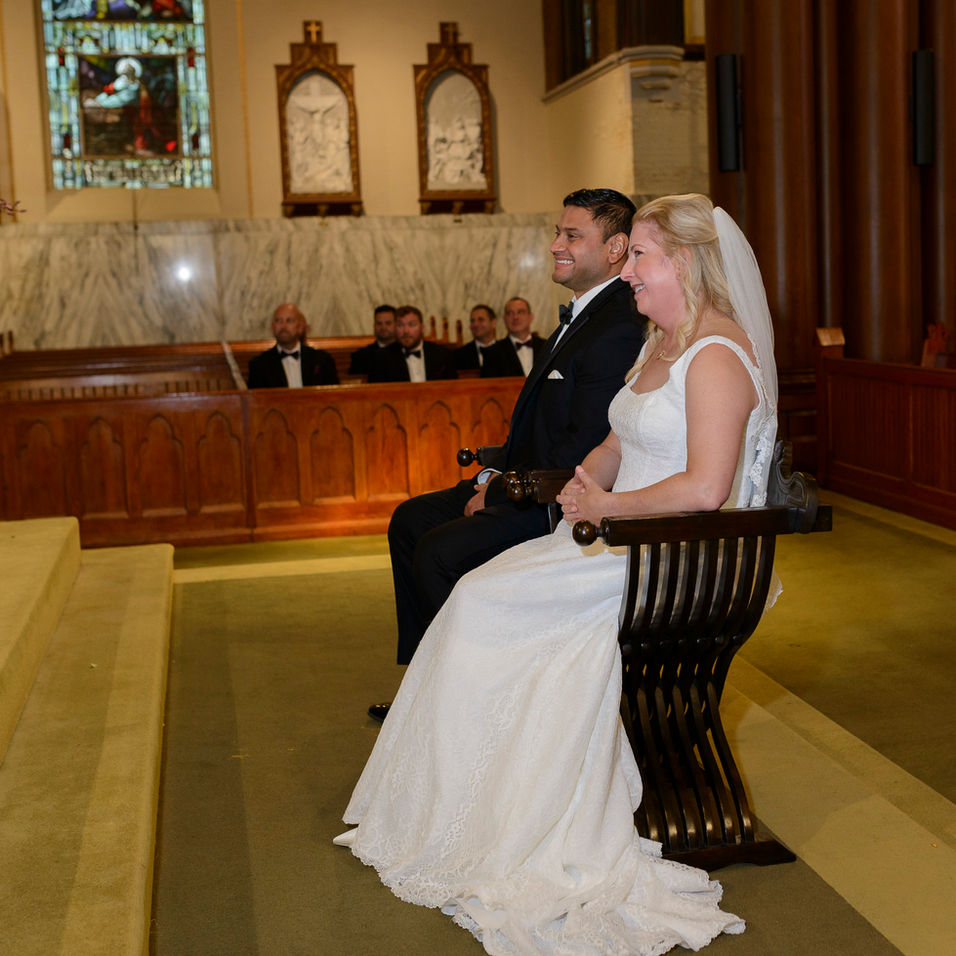 The bride and groom seated side by side on wooden chairs during their wedding ceremony, both smiling joyfully. Behind them, groomsmen in tuxedos sit in the church pews, with ornate stained glass windows and religious artwork adorning the marble wall in the background.