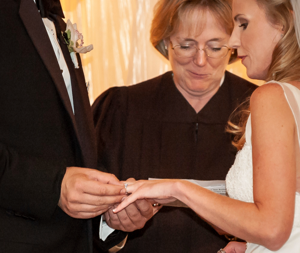 The bride and groom exchange rings during the wedding ceremony, with the groom gently placing a ring on the bride’s finger. The officiant, dressed in a black robe, stands behind them with a warm expression. The moment is illuminated by soft, warm lighting, highlighting the intimacy and significance of the exchange.