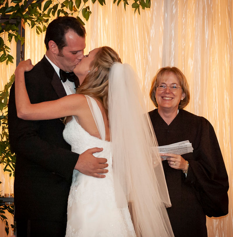 The bride and groom share their first kiss as a married couple, with the bride in a white gown and veil and the groom in a black tuxedo. The officiant stands beside them, smiling warmly and holding ceremony notes. The backdrop of softly lit fabric and greenery enhances the romantic moment.
