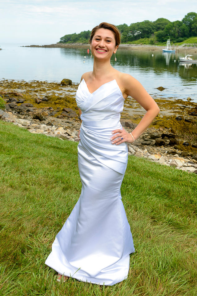 A radiant bride poses outdoors in a stunning white gown with elegant draping, standing on lush green grass by a serene waterfront. The calm water and rocky shoreline provide a picturesque backdrop, complemented by a few boats and distant greenery. The bride smiles brightly, exuding joy and elegance on her special day.
