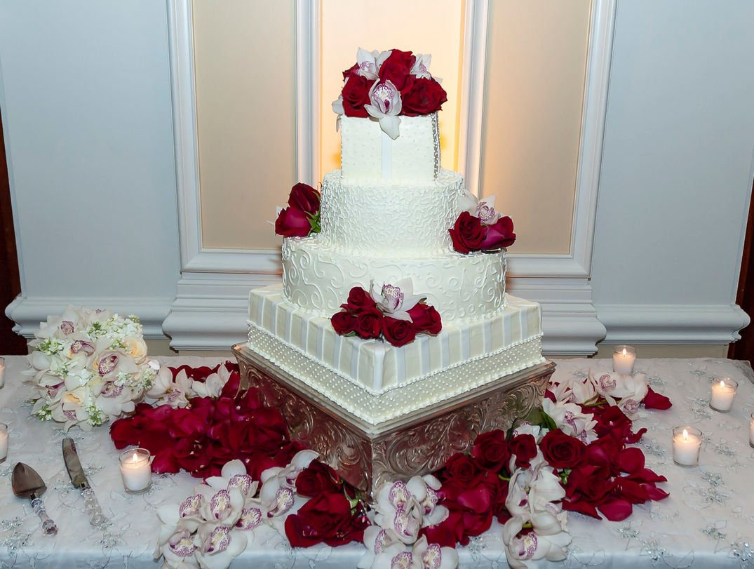 A stunning three-tier wedding cake decorated with intricate white frosting patterns and adorned with vibrant red roses and white orchids. The cake sits on an elegant silver stand, surrounded by a table beautifully arranged with matching flowers, candles, and the bride’s bouquet, creating a romantic and sophisticated display.