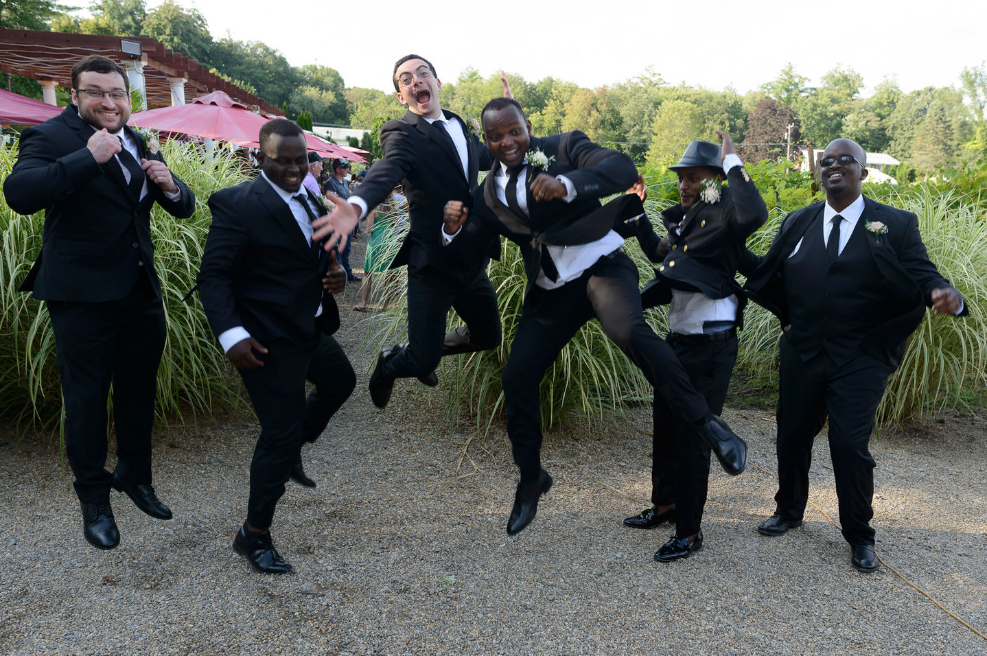 The groom and his groomsmen share a fun, energetic moment as they jump into the air together for a group photo. All are dressed in black suits with ties, some adding playful touches like a hat. The backdrop features lush greenery and pink umbrellas, capturing the lively spirit of the celebration.