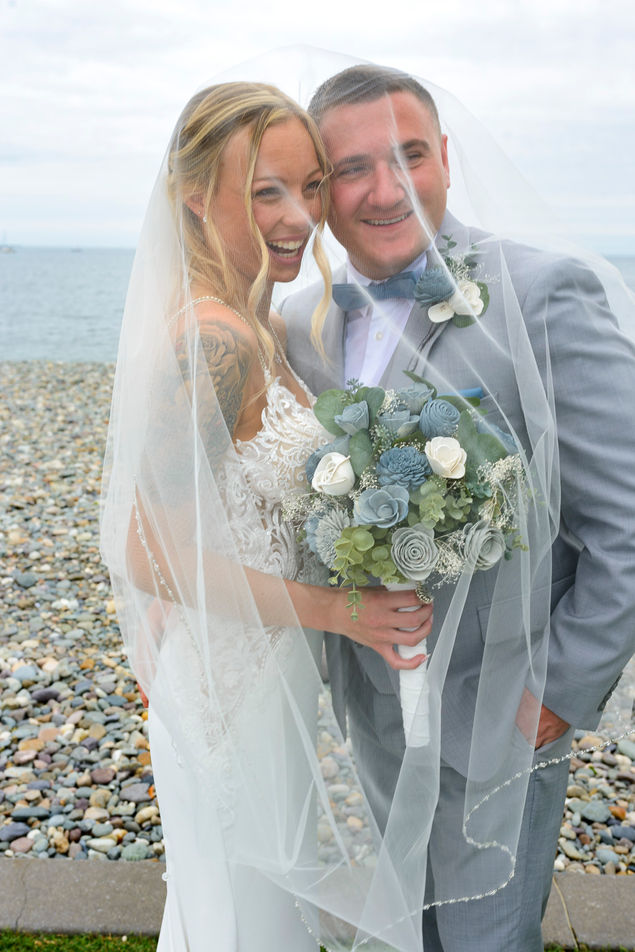 Bride and groom share a happy moment under the bride's veil, with wide smiles on their faces. The groom in a light gray suit holds a bouquet of blue and white roses, matching his blue bow tie, while the bride in a detailed lace wedding gown, her tattoo visible, beams with happiness. They are surrounded by a natural backdrop of a pebble beach and the ocean.