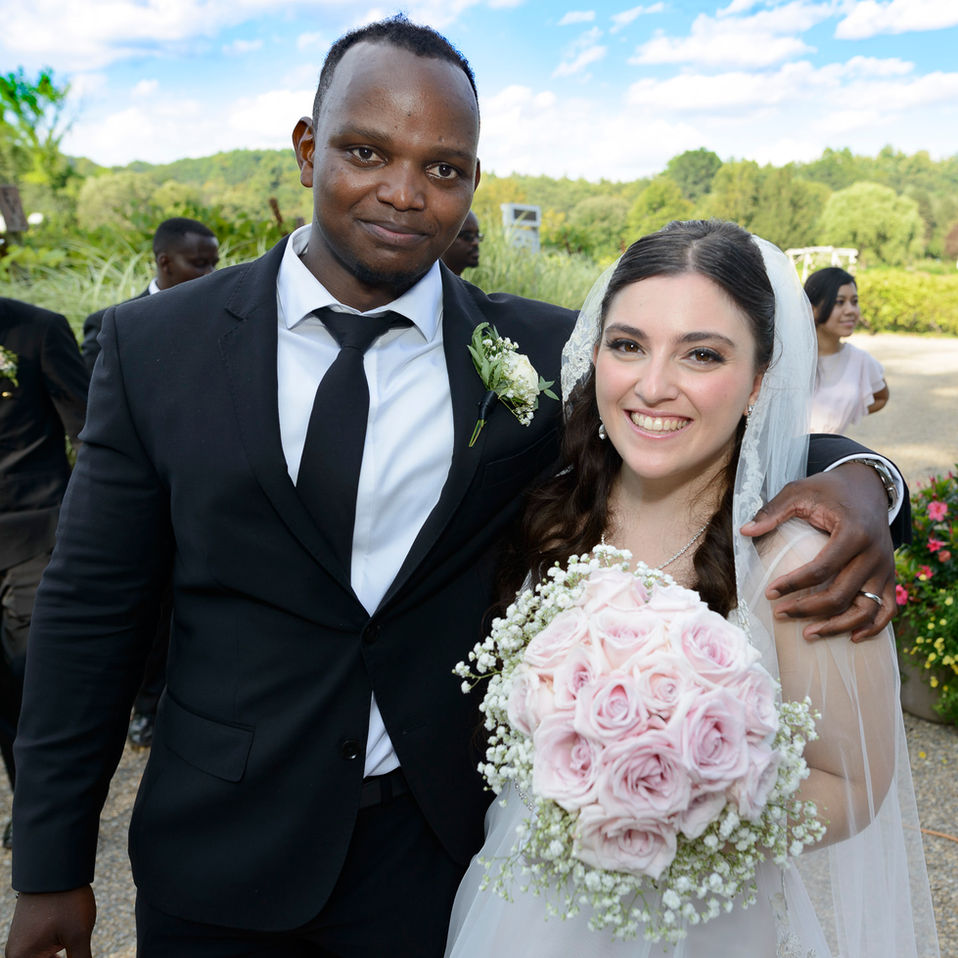 A close-up portrait of the newlyweds outdoors, smiling happily on their wedding day. The groom wears a black suit with a white boutonniere, and the bride is radiant in her lace gown and veil, holding a bouquet of pale pink roses and baby's breath. The background features greenery and a bright blue sky, capturing the joyful moment.