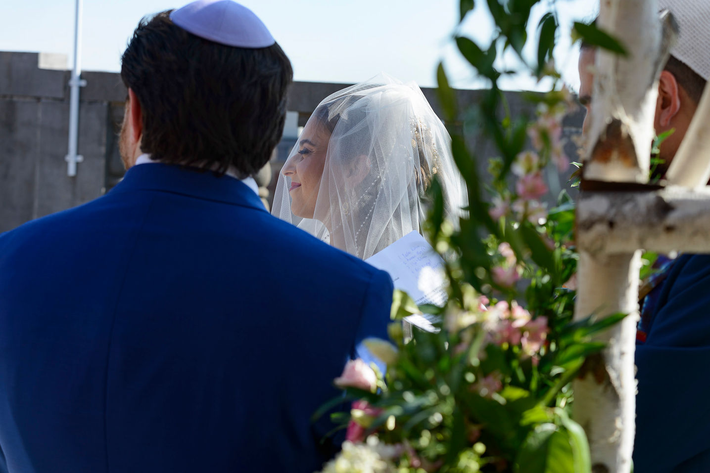 A candid wedding photograph taken during a rooftop ceremony. The bride, wearing a veil and smiling softly, is captured in profile as she exchanges vows with the groom, who is seen from behind wearing a blue suit and kippah. The scene is framed by floral arrangements and a birchwood canopy, adding natural elegance to the intimate moment.