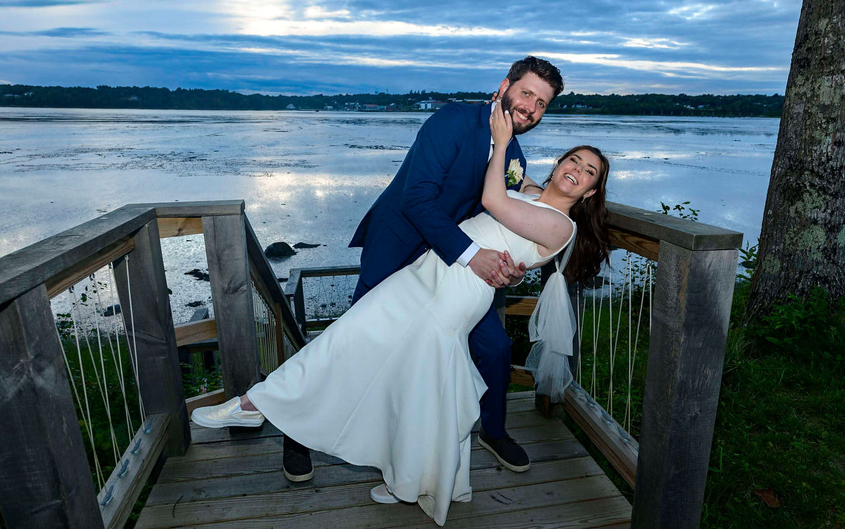 Bride and groom pose in a playful dip on a wooden dock overlooking the water at twilight.
