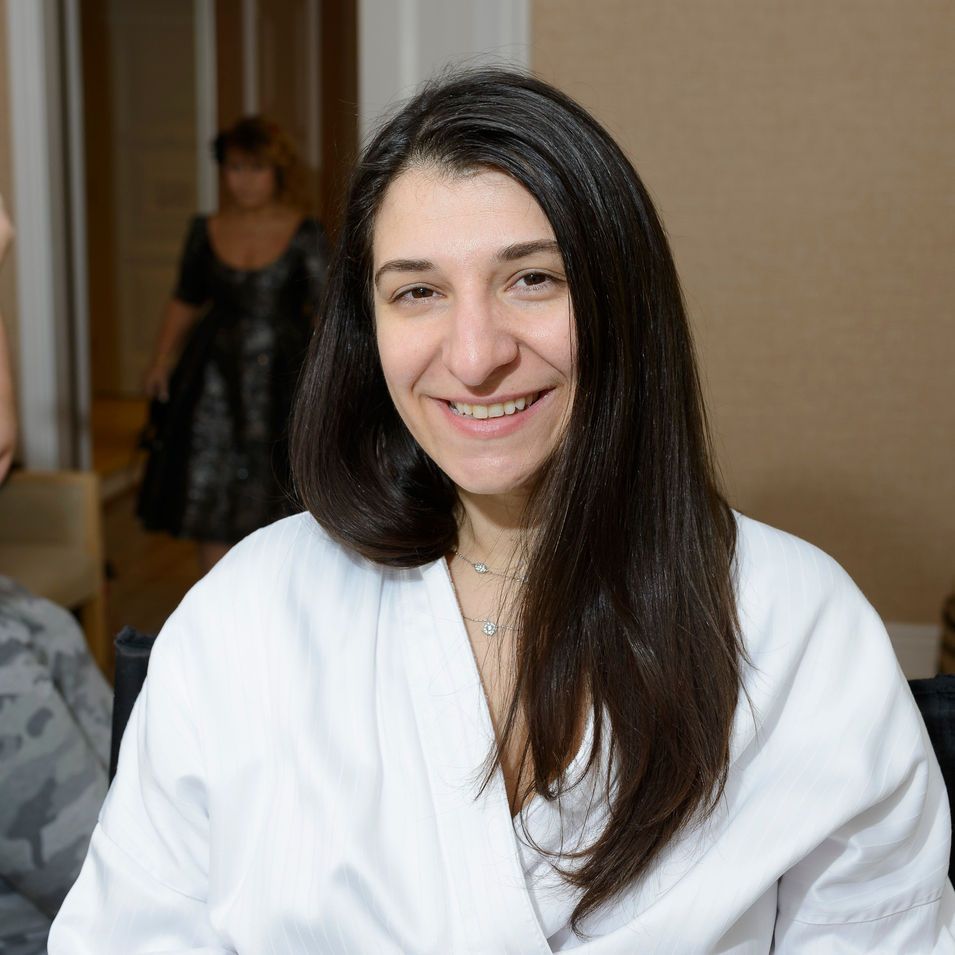 A wedding preparation photograph showing the bride before makeup. She is seated in a white robe with a natural and radiant smile, her long, straight hair framing her face. The background includes a softly blurred figure, adding context to the pre-ceremony setting. The bride's joyful expression captures the excitement and anticipation of her special day.