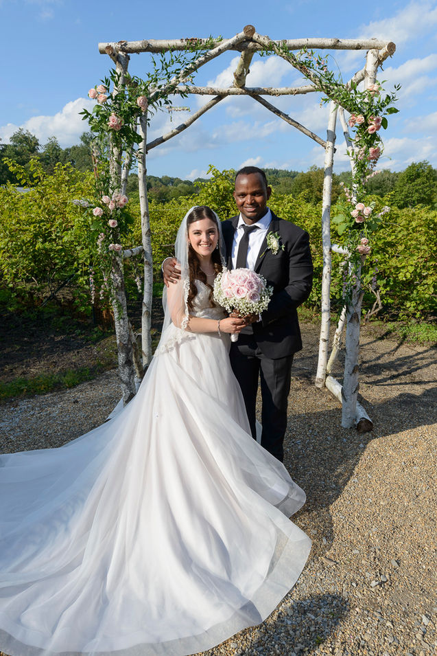 The bride and groom stand together under a birchwood wedding arch adorned with pink roses and greenery, smiling warmly. The bride wears a flowing white gown with a lace veil, holding a bouquet of pale pink roses and baby's breath, while the groom looks sharp in a black suit with a white boutonniere. The backdrop of a sunny vineyard and blue sky adds a romantic touch to the scene.