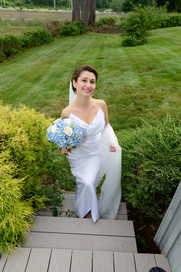A wedding photograph of a smiling bride standing on wooden steps outdoors, holding a bouquet of blue and white flowers. She wears a strapless white gown with a flowing veil, surrounded by lush greenery and a manicured lawn in the background.