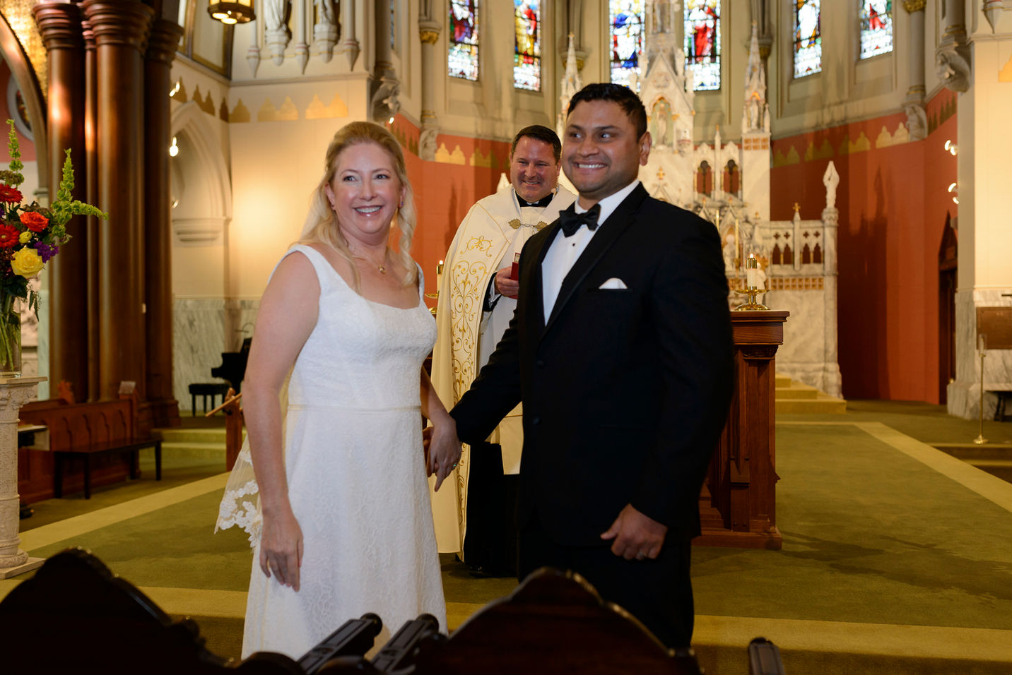 The newlyweds smile brightly as they hold hands at the altar. The bride is dressed in a simple yet elegant white gown with lace details, and the groom wears a sharp black tuxedo with a bow tie. The priest stands behind them, smiling, against the backdrop of the ornate church interior adorned with colorful stained glass windows and floral arrangements.