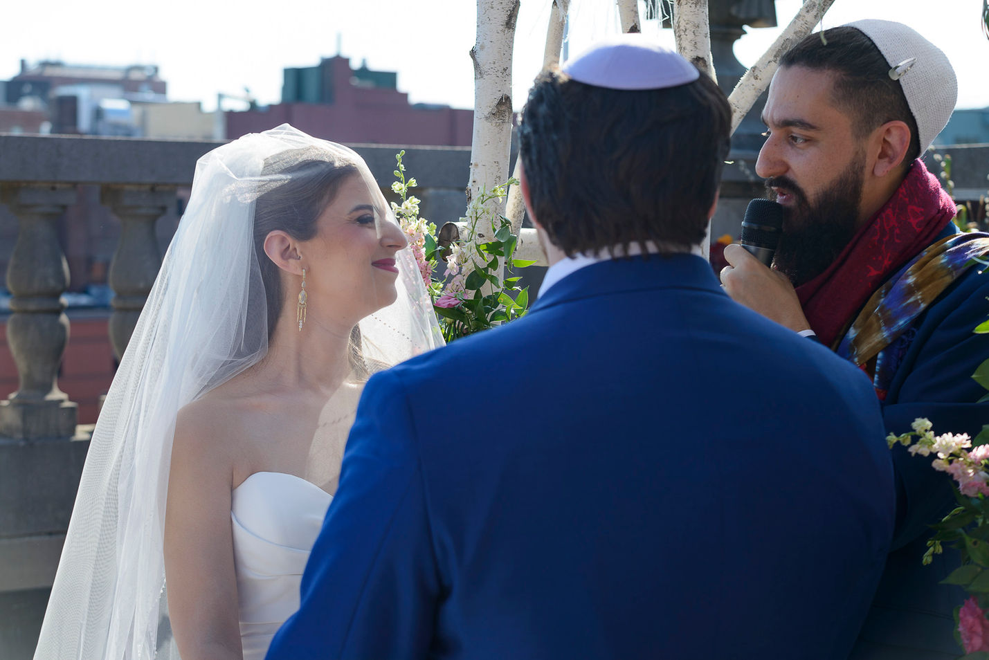 The bride and groom stand together under the chuppah during their rooftop wedding ceremony. The bride, glowing in her white gown and veil, smiles lovingly at her groom, who is dressed in a blue suit and kippah. The officiant, holding a microphone, speaks with heartfelt words, surrounded by soft floral arrangements and the backdrop of an urban skyline.