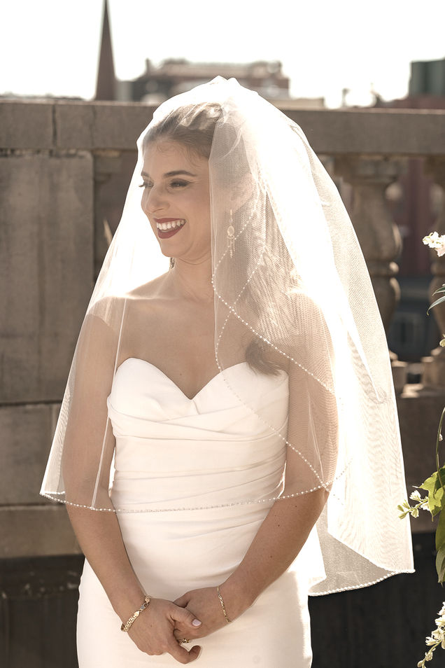 The bride stands at the rooftop ceremony, radiant in her strapless white gown and pearl-trimmed veil. She smiles warmly, her hands clasped in front of her. Sunlight softly illuminates her veil and highlights the elegant details of her bridal look, with the urban skyline in the background.