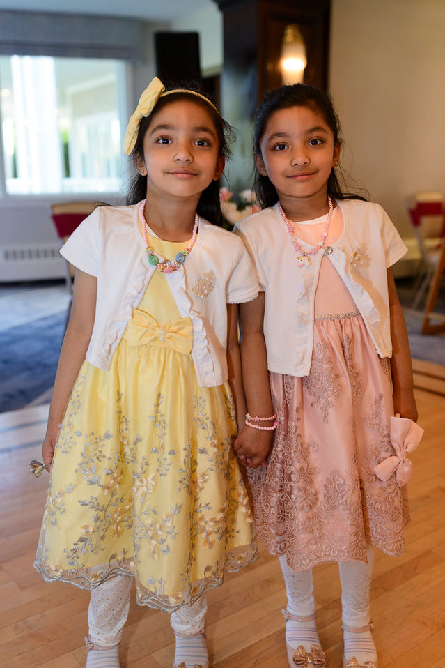Two young girls possibly twins dressed elegantly for a Pakistani wedding celebration. One is wearing a yellow dress with silver floral embroidery, while the other is in a pink dress with intricate lace details. Both are adorned with matching white cardigans, colorful beaded necklaces, and white leggings. They hold hands and smile sweetly, capturing the festive and joyous spirit of the event.