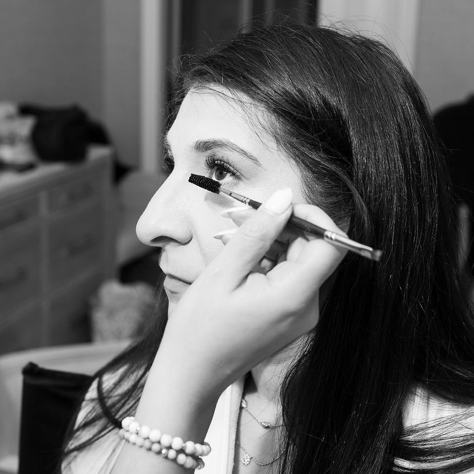 A close-up black and white photo of a bride having mascara applied by a makeup artist. The bride, partially facing the camera, appears calm and reflective with her eyes wide open. The makeup artist's hand is visible, carefully wielding the mascara brush. The bride's jewelry, including a delicate necklace and a pearl bracelet, adds elegance to the serene moment.