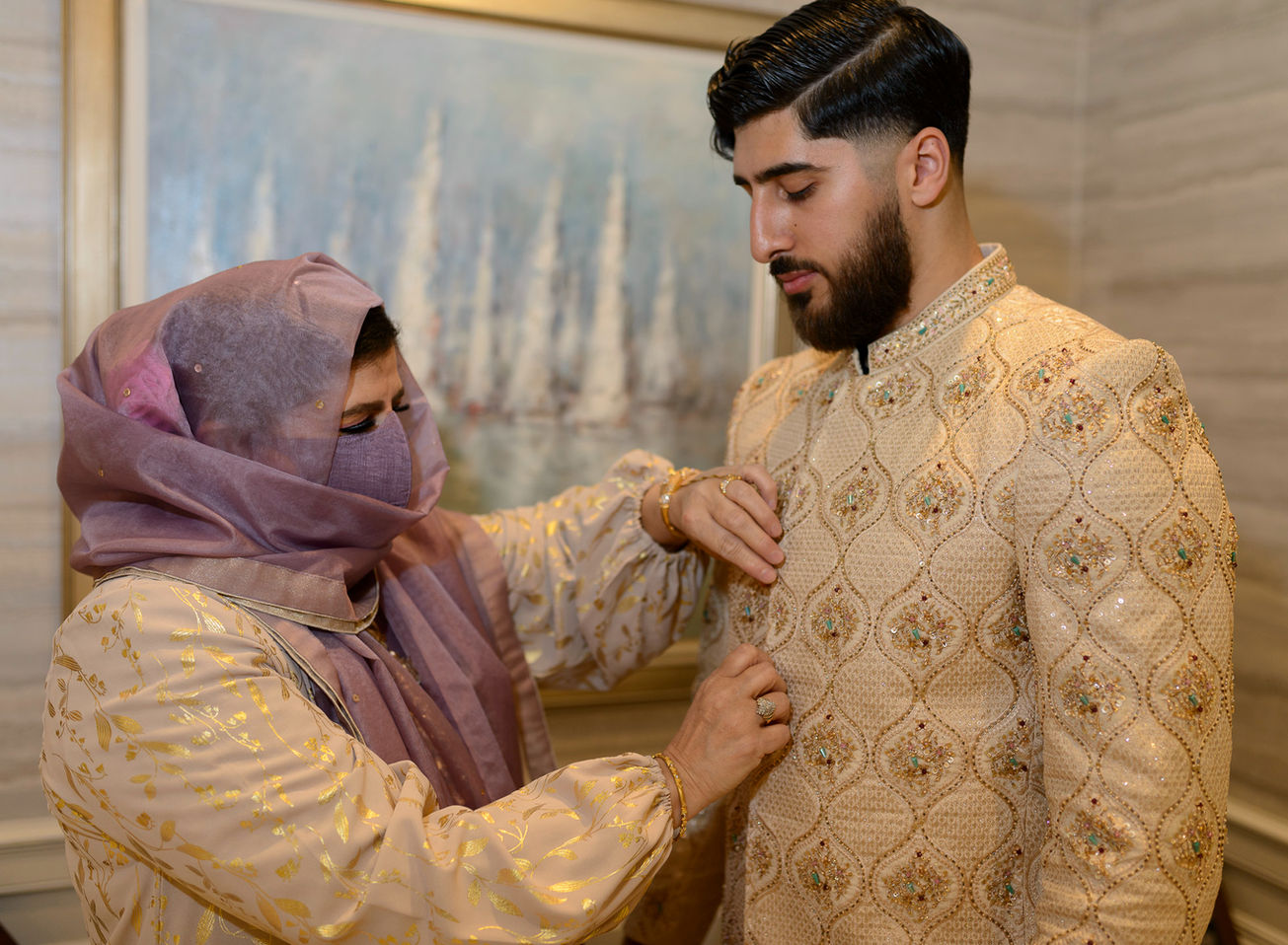 A touching moment at a Pakistani wedding where the mother, dressed in a delicate cream color outfit with golden accents and a lavender scarf covering her head, helps her son adjust his intricately embroidered golden sherwani. The groom looks down attentively, sharing a quiet moment with his mother in a warmly lit room adorned with soft decor.