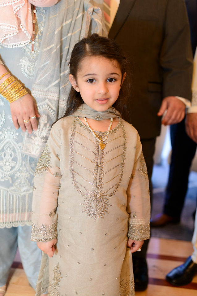 A young girl at a Pakistani wedding celebration wearing a beige traditional outfit embellished with intricate embroidery and paired with a matching dupatta. She is accessorized with a delicate pearl necklace and a gold pendant. The background features elegantly dressed adults, highlighting the festive and joyous atmosphere.