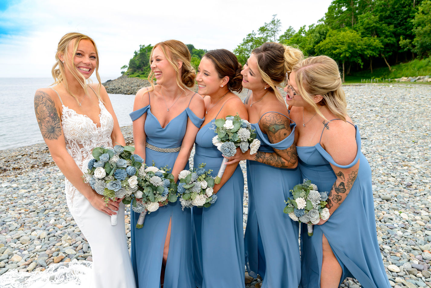 A joyful bride and her bridesmaids pose by the seaside, sharing a laugh. The bride, positioned centrally, is dressed in an elegant white lace gown, while her bridesmaids wear matching cold-shoulder, slate blue dresses. Each holds a bouquet of blue and white flowers, complementing the ocean backdrop and pebbled shore. Their happiness and camaraderie are evident in their bright smiles and close gathering.