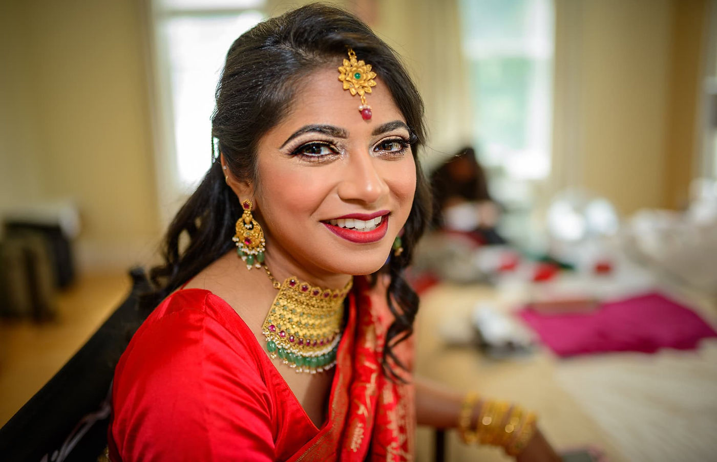 Smiling Bangladeshi bride wearing traditional red bridal attire and gold jewelry while getting ready for her wedding.