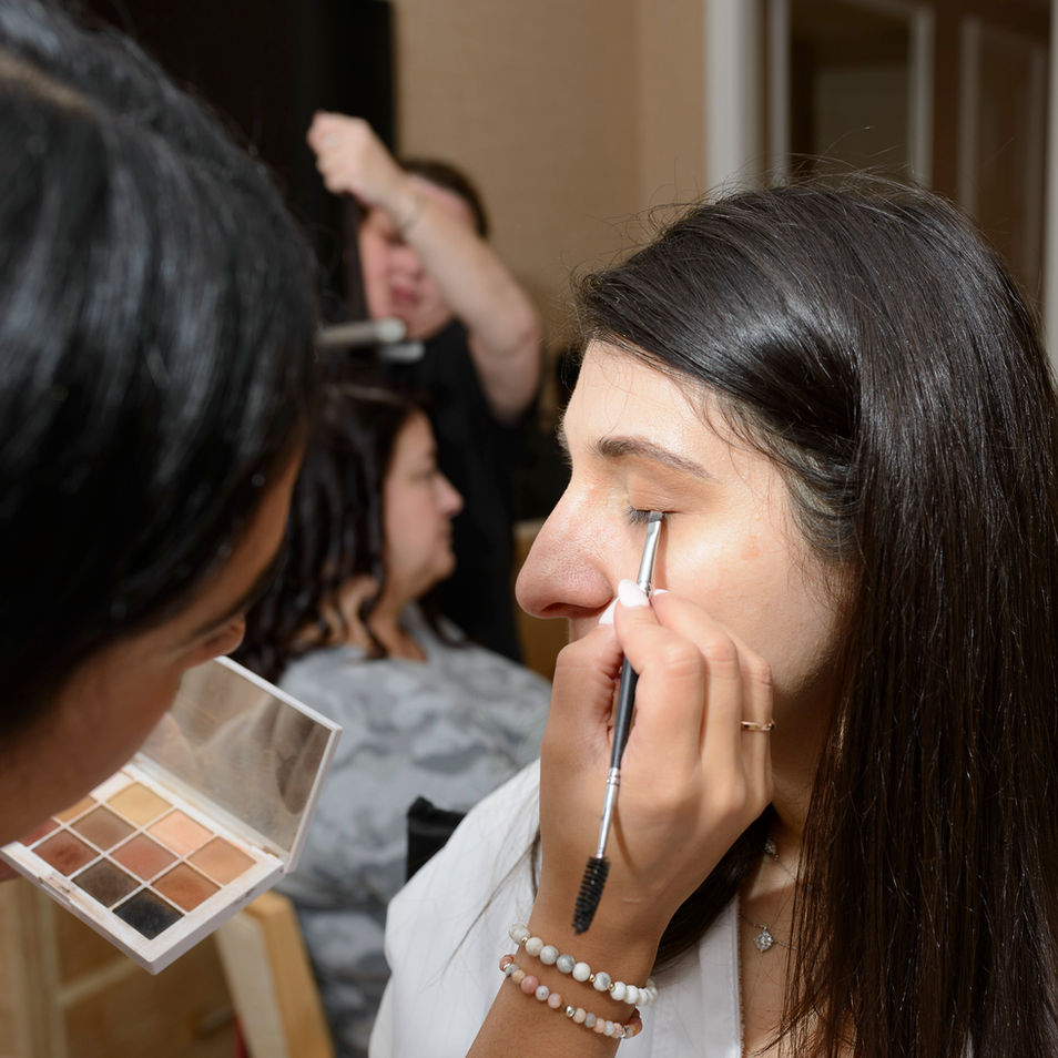 A wonderful wedding preparation photograph showing the bride getting her makeup done. A makeup artist carefully applies eyeliner using a precision brush, while holding a palette of neutral-toned eyeshadows. The bride has her eyes closed, her relaxed expression reflecting the calm before the wedding. In the blurred background, another person is getting their hair styled, adding to the bustling preparation scene. The moment captures the intricate details of getting ready for the special day.