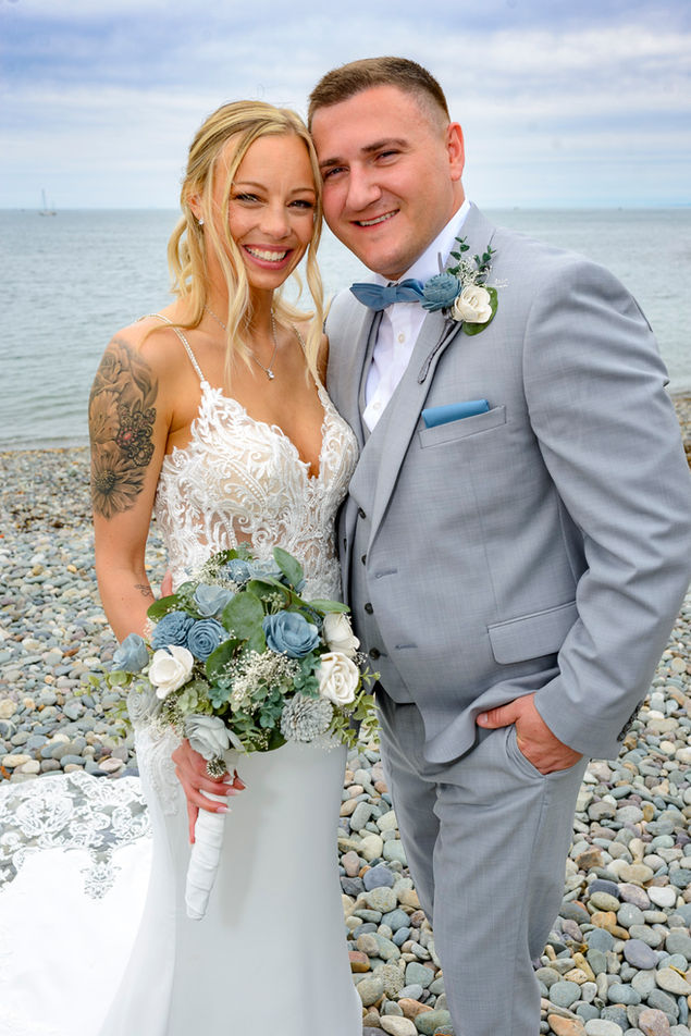 Portrait of a joyful bride and groom standing on a pebbly beach. The bride, with a radiant smile, showcases her detailed lace wedding dress and a full-sleeve tattoo, holding a bouquet of blue and white flowers. The groom, in a light grey suit with a blue bow tie and boutonnière, stands close, sharing a happy moment. The ocean and overcast sky provide a natural, serene backdrop to their celebration.