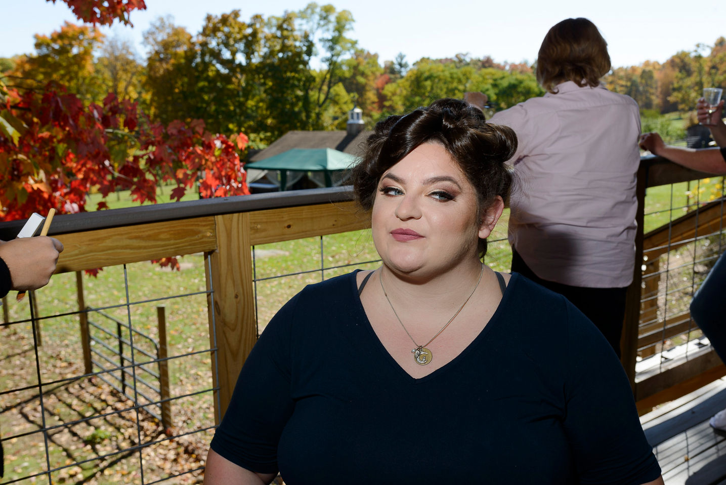 A wedding photograph capturing the bride sitting outdoors on a wooden deck surrounded by vibrant autumn foliage. She has her makeup beautifully done with soft curls pinned up, wearing a navy blue top and a delicate necklace. Guests in the background enjoy the scenic fall view.