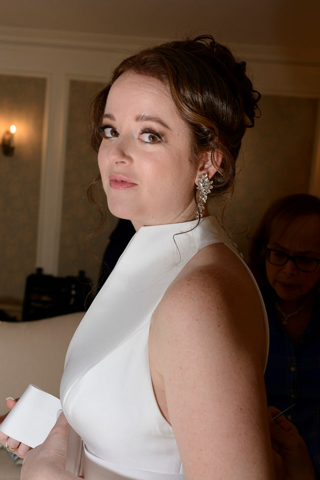 Close-up portrait of a bride in a sophisticated white gown with a high neckline, looking over her shoulder with a soft smile. Her hair is styled in an elegant updo, and she wears ornate floral earrings. In the background, an older woman, possibly a family member, helps with a final touch on the bride's dress, adding a heartwarming detail to the preparation scene.