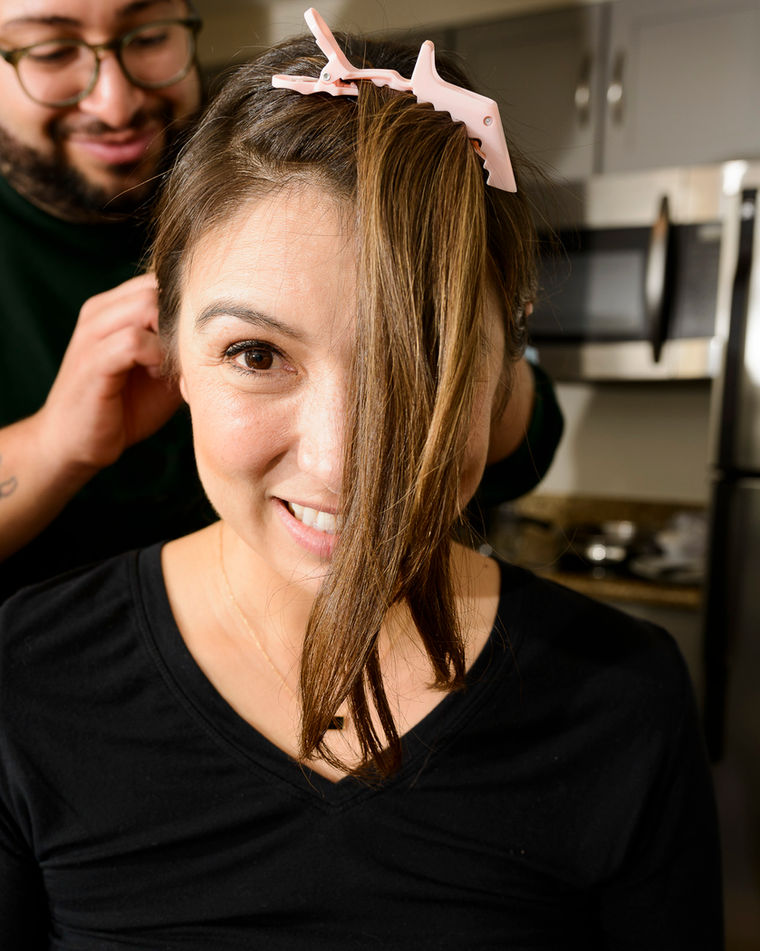 Bridesmaid smiling while having her hair styled, with pink hair clips holding sections of her hair in place. A hairstylist is seen working in the background. The bridesmaid wears a black top and is sitting in a modern kitchen setting.