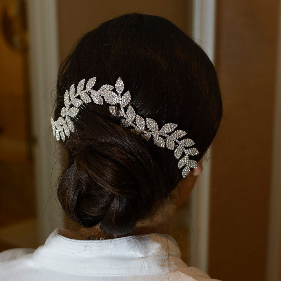 A close-up photograph of the bride's elegant hairstyle, featuring a sleek bun adorned with a sparkling, leaf-shaped rhinestone hair accessory. The intricate details of the accessory and the smooth finish of the hairstyle create a sophisticated and timeless look for the special day.