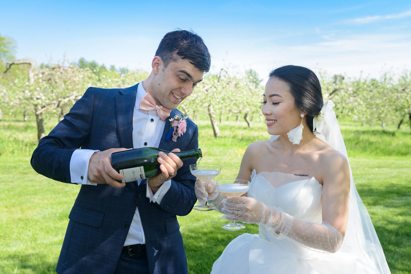 Groom pouring champagne into the bride's glass as they celebrate outdoors in an apple orchard. The groom wears a navy suit with a pink bow tie and boutonniere, while the bride, dressed in a strapless white gown and sheer polka-dot gloves, smiles warmly. The sunny setting and lush green orchard create a joyful and romantic atmosphere.