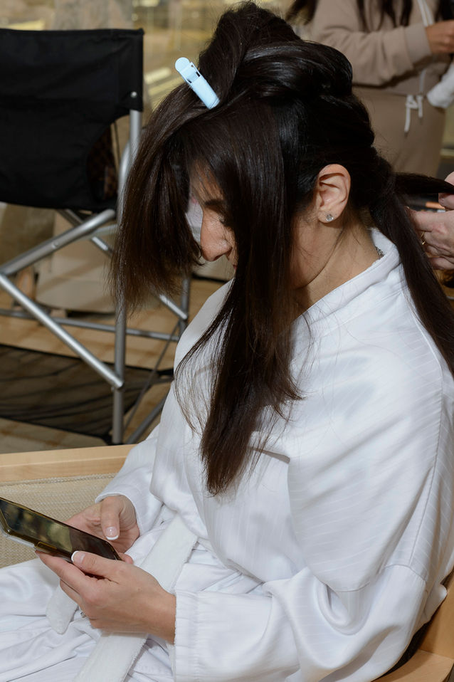 A bride is seated, looking down at her smartphone while her hair is styled in preparation for her wedding. She wears a white robe with her long, dark hair partially pinned up, featuring a white clip holding sections in place. The background is softly blurred, emphasizing the intimate and quiet moment in a bustling preparation room.