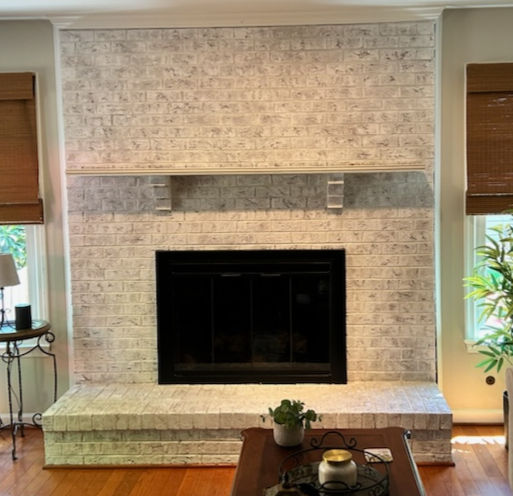 White brick fireplace with a black metal insert, wooden blinds on windows, hardwood floor, and a plant on the wooden table. Cozy setting.