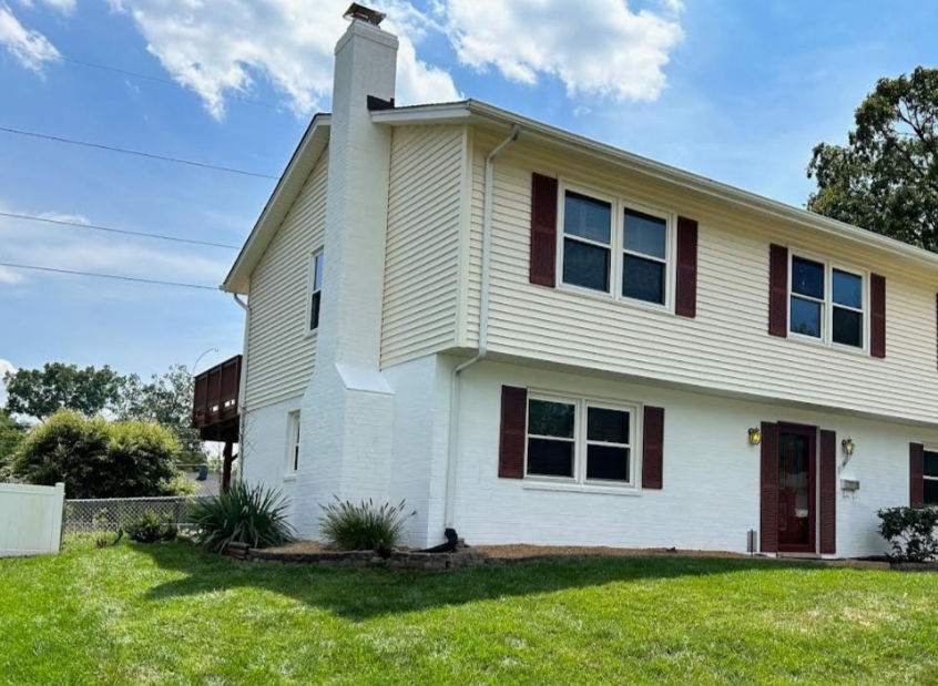 Two-story white house with maroon shutters, chimney, and green lawn. Blue sky with clouds in the background. Peaceful suburban setting.