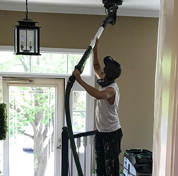 Worker using a vacuum to clean a high ceiling in a well-lit room with large windows. Paint-splattered clothes indicate home renovation.