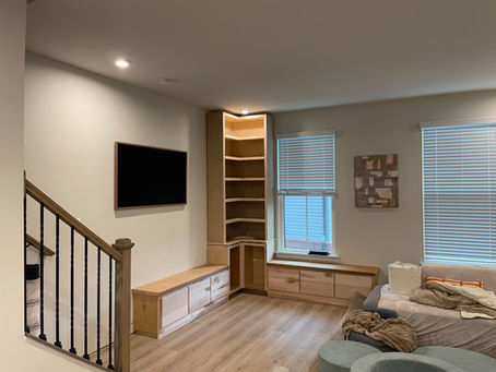 Modern living room with a cozy sofa, blankets, and a laundry basket. Wood shelves, TV mount, art on wall, and large windows with blinds.