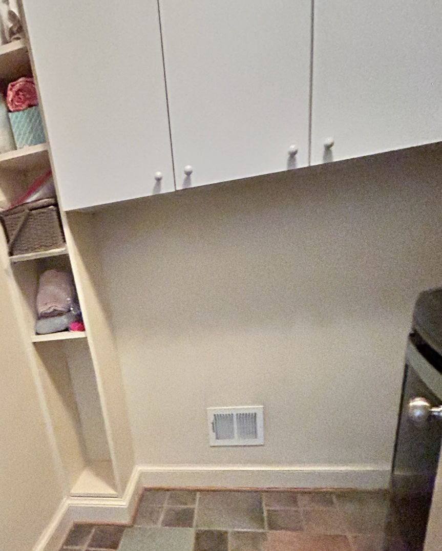 White cabinets and shelf with folded clothes and baskets in a tiled room. Beige walls, floor vent, and washer visible, creating an organized look.